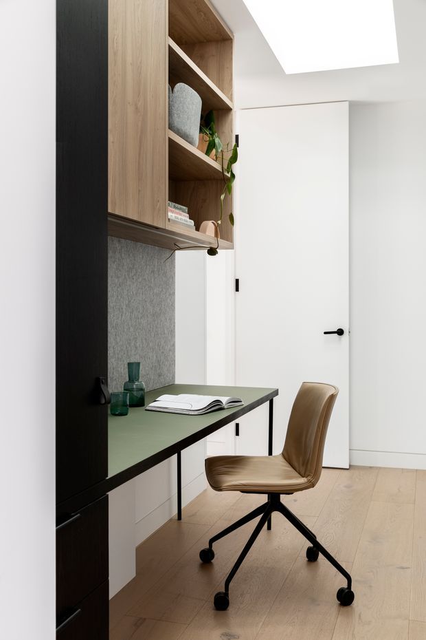 A compact, contemporary home office nook with a built-in desk, green worktop, white cabinetry, open timber shelving, and a tan upholstered swivel chair. The space has pale timber flooring, a grey textured backsplash panel, black joinery accents, and a large ceiling skylight that provides bright natural light.