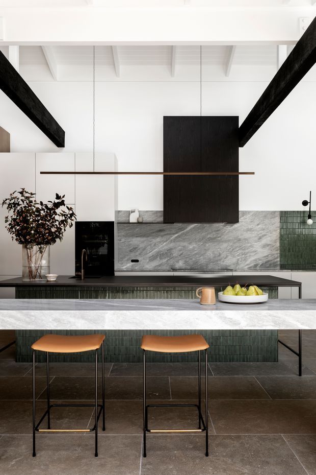 A refined contemporary kitchen with a large stone island and integrated dining overhang, slim black-framed bar stools, dark green vertically ribbed cabinetry, and a matching green tiled feature wall. The room has a restrained palette of white, grey stone, charcoal, black timber beams, and warm tan upholstery. Exposed white ceiling rafters and bold black structural beams add architectural character, while a linear pendant light spans the island.