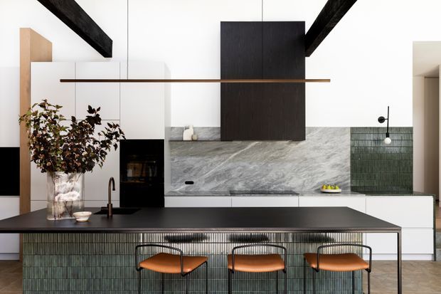 Contemporary open-plan kitchen with a large dark island, integrated seating, veined stone backsplash, dark timber cabinetry, green glazed-tile wall sections, and exposed black ceiling beams. Warm timber and leather accents contrast with the monochromatic finishes.