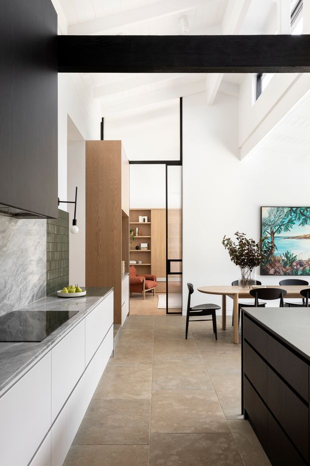 A bright open-plan kitchen and dining area with a double-height white interior and exposed white-painted beams. The kitchen features white flat-panel cabinetry, a dark stone benchtop, green tile splashback, and a large black island. A timber partition and shelving divide the kitchen from a warm-toned living area, while black-framed glazing and furniture provide strong contrast. Large beige stone-look floor tiles and a coastal artwork complete the space.