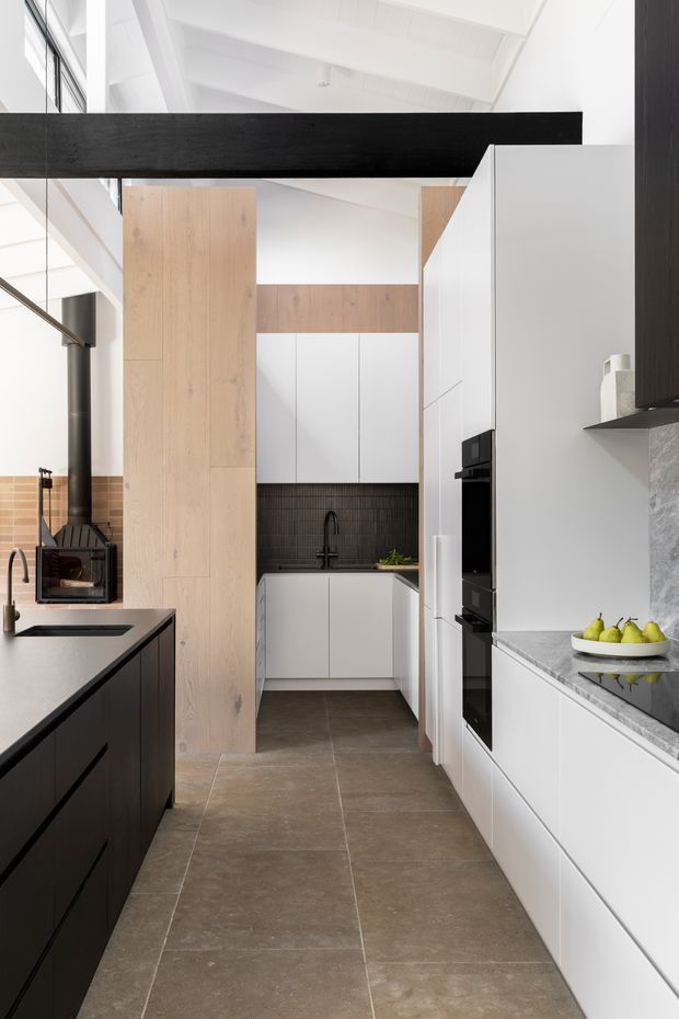 A bright, contemporary galley kitchen with a tall white-painted ceiling and exposed dark timber beam. The space features white handleless cabinetry, pale timber vertical joinery, dark textured tiled splashback, large taupe-brown floor tiles, and grey stone benchtops. A black wood-burning stove sits beside the island, while integrated black ovens and a fruit bowl occupy the opposite cabinetry run.