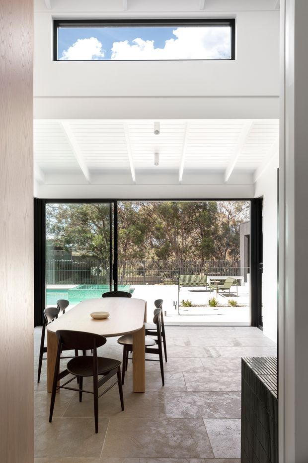 Bright open-plan dining area opening directly onto an outdoor terrace and swimming pool. The room features a large timber dining table with dark chairs, pale large-format tiled flooring, black-framed full-height sliding glass doors, white walls, and a distinctive white slatted ceiling with exposed beams. A high horizontal clerestory window brings in additional daylight, while trees and a pool are visible outside.