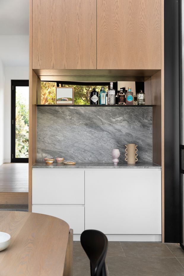 Contemporary built-in dining room cabinetry featuring warm timber overhead cupboards, white lower drawers and cupboards, and a dramatic grey veined stone display backsplash and benchtop. An open shelf displays bottles and decorative objects, with a timber dining table and black chair in the foreground. Dark-framed glazing and a timber floor are visible to the side.