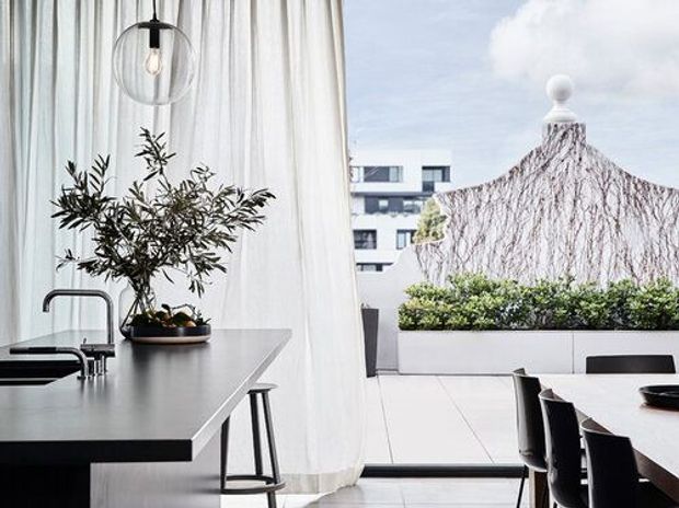 Contemporary dining and kitchen interior opening onto a bright rooftop terrace or balcony. The image features a dark stone or composite kitchen island with integrated sink and minimalist tapware, a sculptural glass pendant, sheer white curtains, and a dining table with black chairs. Large glazing provides views of the terrace, planted screening, and adjacent urban buildings.