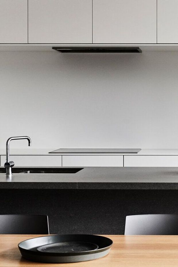 A contemporary kitchen viewed across a timber dining table, featuring a dark stone island benchtop with an integrated sink and minimalist chrome tap, white handleless cabinetry, a black linear rangehood, and a pale stone or glass splashback. Black and white dining chairs and a black serving platter add contrast.