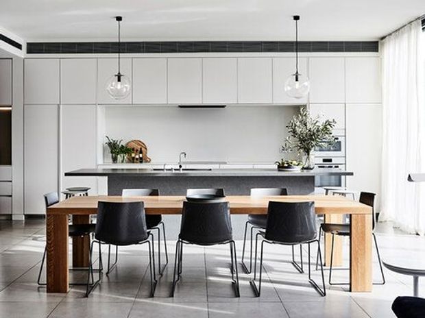 Bright contemporary open-plan kitchen and dining area with floor-to-ceiling white cabinetry, a long island with dark stone benchtop, integrated appliances, pendant lights, and a timber dining table surrounded by black chairs. Large tiled flooring and sheer curtains create an airy, refined interior.