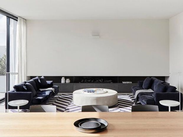 A bright, minimalist open-plan living room with two dark navy sofas facing each other across a large round white coffee table. A long black built-in fireplace or media wall runs along the back beneath a plain white wall, while a bold black-and-white geometric rug anchors the seating area. Floor-to-ceiling glazing with sheer curtains is visible at left, and a timber dining table with a black serving bowl occupies the foreground.