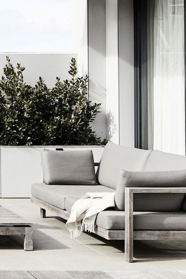 A bright minimalist outdoor lounge area with a low-profile grey sectional sofa and matching armchair arranged on a pale tiled terrace. A white rendered wall, raised planter filled with dense greenery, and tall floor-to-ceiling glazing with sheer curtains form the backdrop. A small weathered timber coffee table and a light throw add subtle texture.