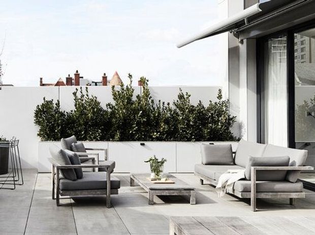 A contemporary rooftop terrace or balcony with grey outdoor lounge furniture arranged around a low rectangular coffee table and recessed fire pit. Large-format grey paving, white built-in planter cabinetry filled with dense greenery, and a dark-framed glazed opening define the space. A retractable awning provides shade, while the urban skyline is visible beyond the terrace wall.