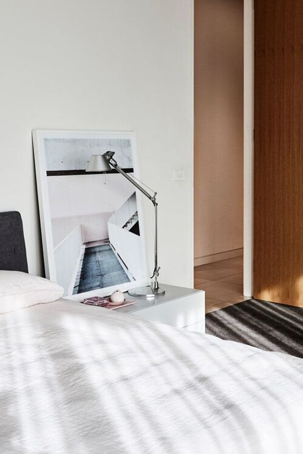 Bright contemporary bedroom featuring a white bed with crisp bedding, a dark upholstered headboard, a minimalist white bedside table, and a silver adjustable task lamp. A large framed architectural artwork leans against the wall, while a warm timber door and grey striped rug add contrast. Sunlight casts soft linear shadows across the bed.