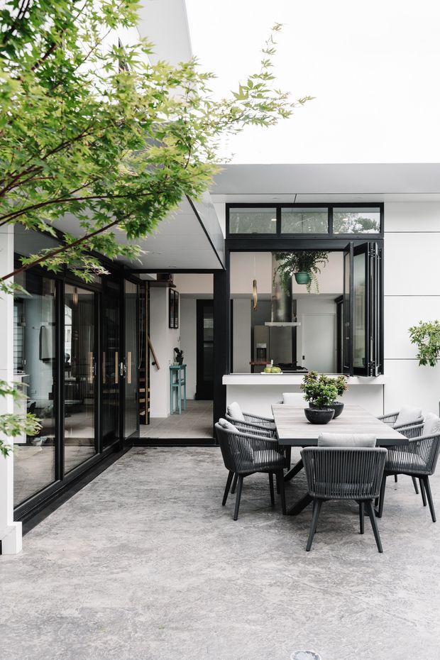 A contemporary courtyard entertaining area adjoining a modern home. The space features a large pale concrete-paved floor, black-framed floor-to-ceiling glazing and folding doors, white horizontal wall cladding, and a servery window opening to the kitchen. A dark outdoor dining table with woven charcoal chairs sits beneath a generous white ceiling, while established leafy planting softens the courtyard.