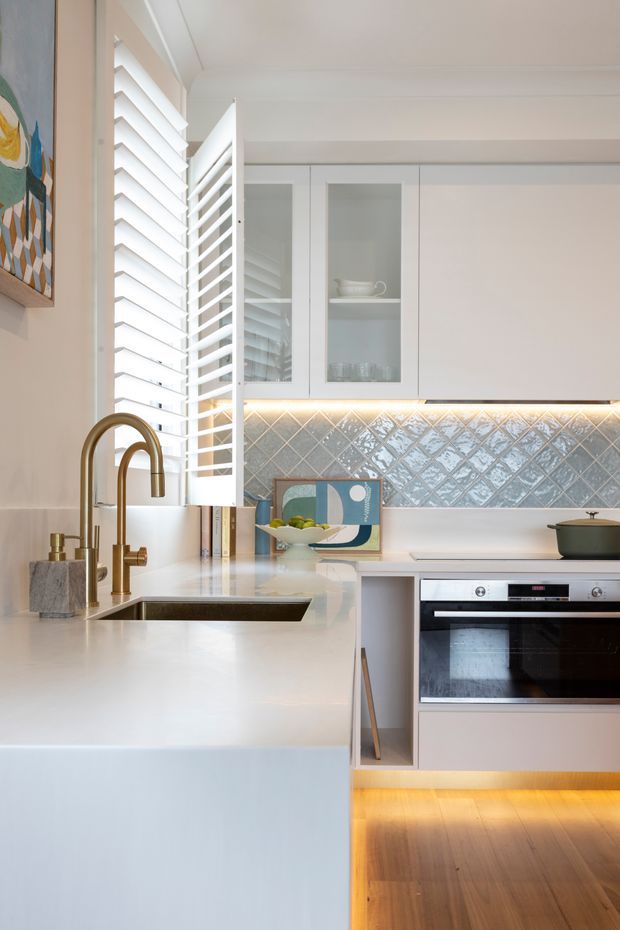 Bright contemporary kitchen with white cabinetry and a large white island featuring an inset sink and brushed brass gooseneck tap. A glossy blue-grey diamond-pattern tiled splashback runs beneath illuminated upper cabinets with glass-front doors. White plantation shutters provide strong daylight, while warm under-cabinet and toe-kick lighting highlights the timber floor. A built-in oven and dark cookware are visible, with restrained artwork and accessories adding colour.