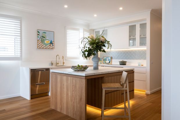 Bright, refined kitchen interior with a central timber-clad island, white cabinetry, glass-front upper cupboards, integrated under-cabinet lighting, timber flooring, and white plantation shutters. A single bar stool sits at the island, while a floral arrangement, artwork, and small countertop accessories add warmth.