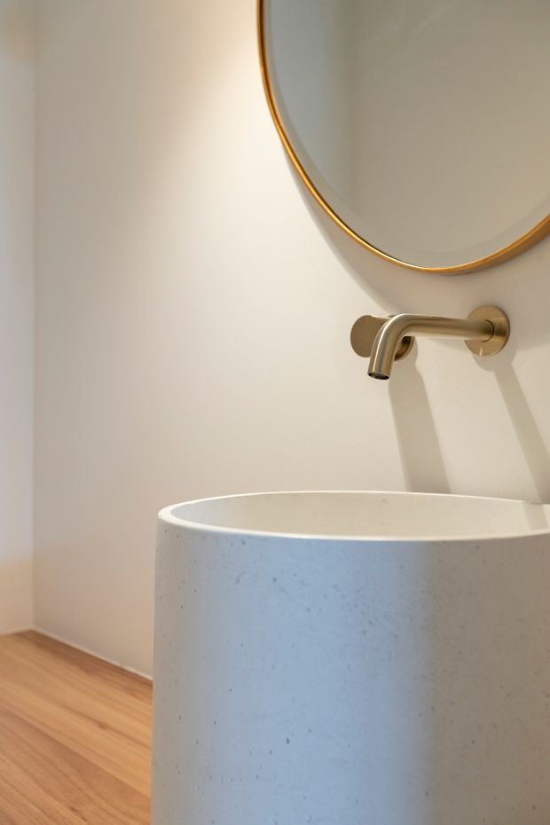 Close-up of a refined bathroom vanity featuring a freestanding cylindrical vessel basin in a pale speckled finish, a wall-mounted brushed brass tap, and a large round mirror with a slim brass rim. Warm timber flooring and soft off-white walls create a calm, minimalist interior.