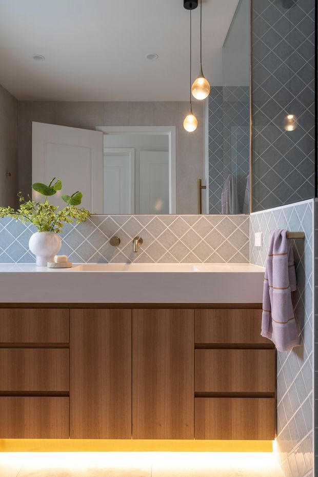 Contemporary bathroom vanity featuring a wide floating timber-grain cabinet with integrated drawers, a pale stone or solid-surface benchtop, and grey-blue diamond-pattern ceramic wall tiles. A large mirrored cabinet spans the vanity, with warm brass-toned tapware and two globe pendant lights. Soft under-vanity lighting and lavender towels add warmth and contrast.