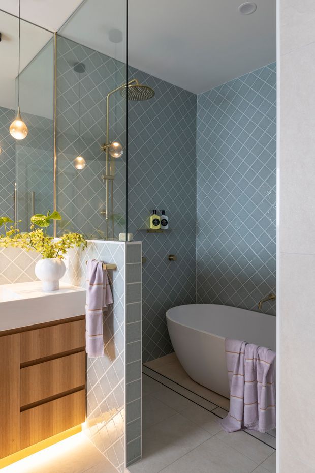 A refined contemporary bathroom with a freestanding white bathtub, walk-in shower, wall-mounted shower fixtures, and a timber vanity with integrated under-cabinet lighting. Blue-grey geometric wall tiles form a diamond pattern throughout the wet area, complemented by pale floor tiles, frameless glass shower screening, and brass-toned fixtures. A large mirrored wall reflects pendant lighting and greenery, while soft lilac towels add a subtle accent.
