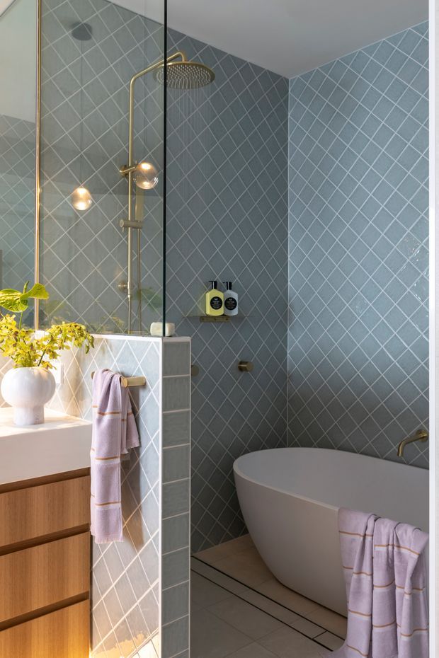 A refined contemporary bathroom with a freestanding white bathtub, glass-enclosed shower, wall-mounted rainfall showerhead, and brass fixtures. The walls and shower partition are clad in pale blue-grey diamond-pattern ceramic tiles with contrasting light grout. A warm timber vanity with integrated basin sits at left, accompanied by a rounded white vessel and greenery. Soft lavender towels with subtle stripes add colour, while natural light creates a calm, spa-like atmosphere.