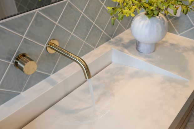 Close-up of a refined bathroom wash area featuring a wall-mounted brushed brass tap flowing over a white integrated basin or countertop. The backsplash is clad in dark grey-green diamond-set tiles with light grout, while the countertop has a soft veined white stone-like finish. A rounded white vase with greenery adds a decorative accent.
