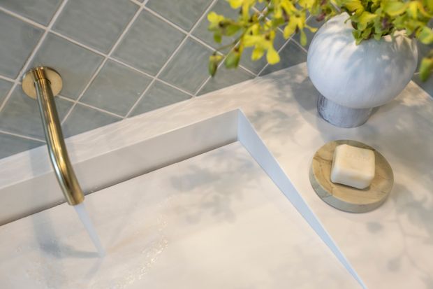 Close-up of a refined bathroom vanity or basin area, showing a white rectangular integrated basin with a slim brass wall-mounted faucet, grey tiled splashback, and a round soap dish beside a decorative vase. The composition is bright and softly styled with warm metallic detailing and neutral finishes.