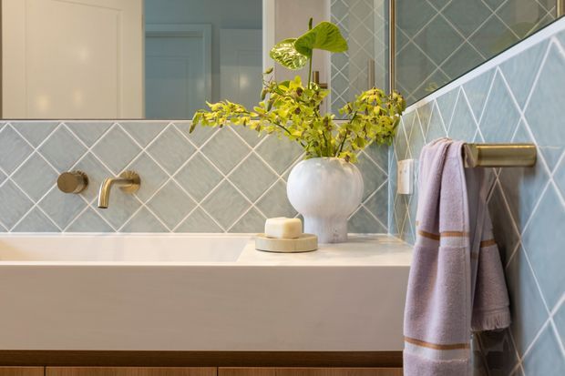 A softly lit contemporary bathroom featuring a built-in bathtub with pale stone or solid-surface surround, blue-grey diamond-pattern wall tiles with light grout, and brushed brass wall-mounted tapware and towel rail. A pale timber vanity is visible below the bath edge, while a white vessel and leafy branch add a natural decorative touch. The overall scheme is refined, calm, and lightly retro-inspired.
