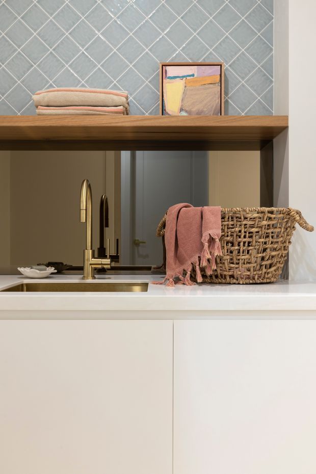 Close-up view of a refined kitchen or kitchenette sink area. The space features pale blue-grey diamond-pattern tiled wall cladding, a warm timber open shelf, white cabinetry and a white benchtop, with a brass-toned mixer tap and undermount sink. A woven basket with a pink towel, folded linens, and a small framed artwork add warm decorative accents. A doorway is visible in the background.