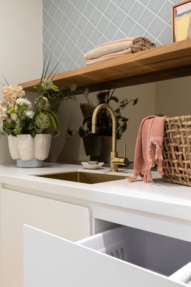 Close-up of a refined laundry or utility-room sink area with a white benchtop, undermount sink, brass gooseneck tap, pale cabinetry and an open drawer. A timber open shelf holds folded towels, while woven storage, a pink towel and botanical arrangements add warmth. The wall features a grey-blue diamond-pattern tiled splashback, with a mirror reflecting greenery and the tap area.
