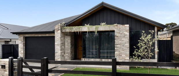 Street-facing exterior view of a contemporary single-storey house with a steep gabled roof, dark vertical cladding, pale brick masonry, large glazed openings, and a projecting balcony or roof terrace with trailing plants. A dark garage door and timber-style boundary fence are visible in the foreground.