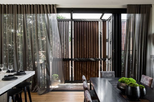 Contemporary dining area with a long dark timber dining table, upholstered dining chairs, and a white island or side counter with stools. Full-height sheer and pleated curtains frame black-framed glazing and a timber slatted privacy screen, creating a strong connection to the outdoor area. Light timber flooring and dark furnishings give the room a refined, moody atmosphere.