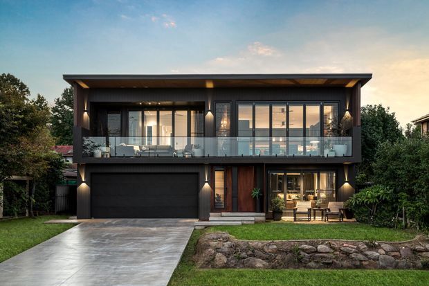 A contemporary two-storey residential facade photographed at dusk, featuring dark horizontal cladding, a broad flat roof with deep eaves, extensive upper-level glazing and a balcony with clear glass balustrades. The ground floor includes an integrated dark garage, timber-toned entry door, and a warmly lit living or dining area opening toward the front garden. A concrete driveway, lawn, landscaped rock retaining edge, and mature planting frame the house.