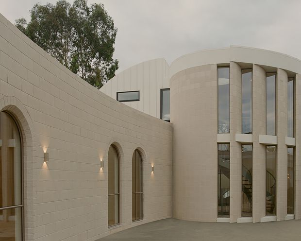 Exterior architectural view of a pale cream masonry building arranged around a paved courtyard. A curved rendered or masonry wall features repeated tall arched openings and discreet wall lights, while the main rounded volume has tall vertical glazed openings separated by substantial curved columns and a smooth curved roofline. The composition is restrained, monumental, and softly lit under an overcast sky.