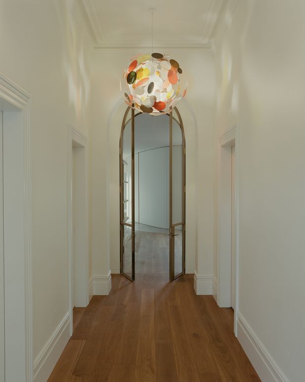 A long, narrow residential hallway with warm timber flooring, pale painted walls, ornate skirting boards and ceiling mouldings, and multiple doorways. At the end is a distinctive tall arched metal-framed glazed partition or doorway, topped by a sculptural circular pendant light with multicoloured translucent elements. The space combines classical architectural detailing with a dramatic contemporary feature.