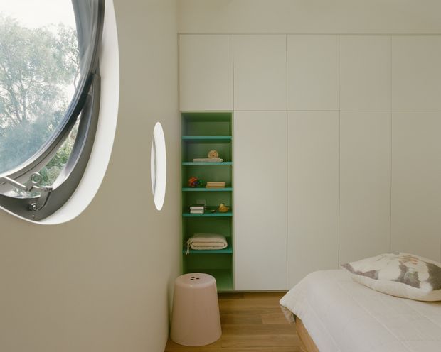 A calm, contemporary bedroom with a low bed at right, white bedding, warm timber flooring, and full-height pale built-in wall cabinetry. A narrow recessed green shelving unit provides a colourful feature, while a sculptural pale stool sits beneath it. The room is illuminated by a large distinctive oval porthole-style window with a dark metal frame.