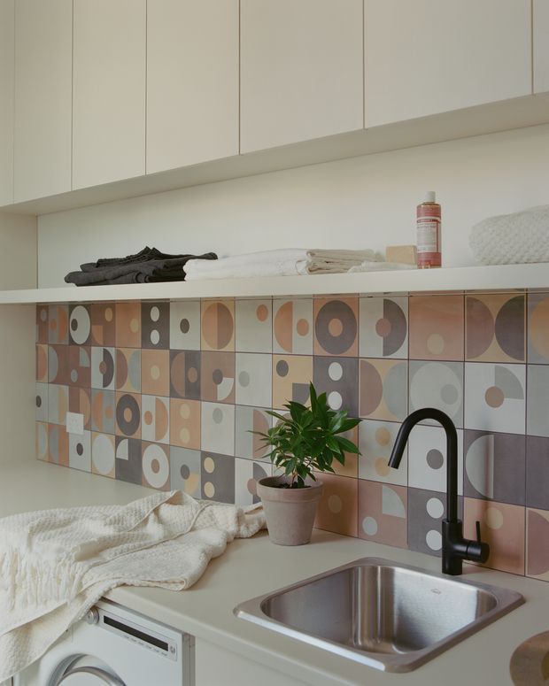 Compact, softly lit laundry room with pale built-in overhead cabinetry, an open display shelf holding folded towels and toiletries, and a countertop with an undermount stainless-steel sink and matte black gooseneck tap. A colourful geometric patterned tile splashback in muted terracotta, beige, cream and grey forms the main feature. A potted plant and folded textured towels add warmth.