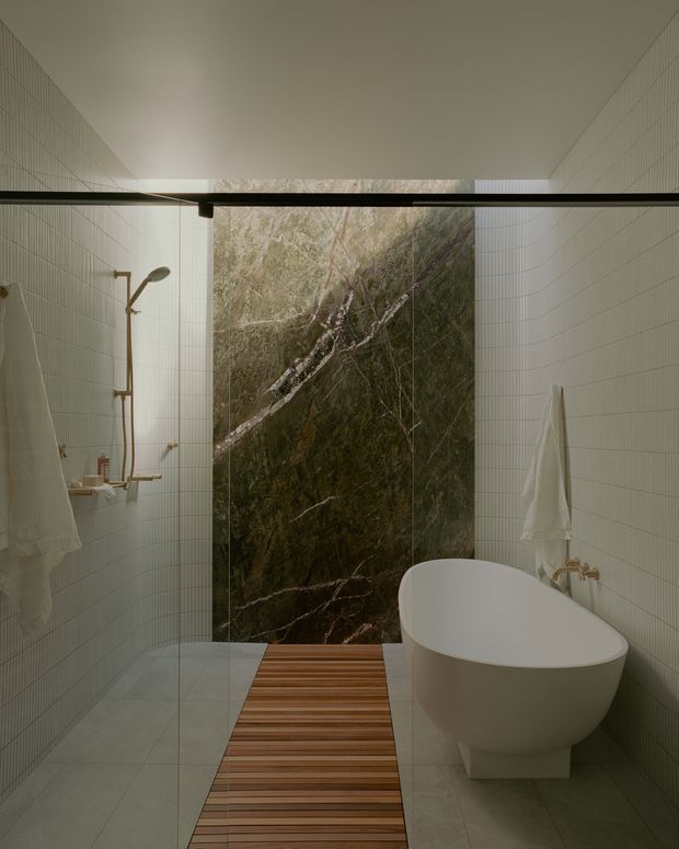A refined modern bathroom with a freestanding white oval bathtub positioned beside a walk-in shower. The shower and surrounding walls are clad in small pale square tiles, while a dramatic dark green-grey veined stone feature wall anchors the rear of the room. A timber slatted bath mat or walkway adds warmth over the pale tiled floor. Brushed brass shower and bath fittings, white towels, and a frameless glass shower screen complete the calm, spa-like composition.