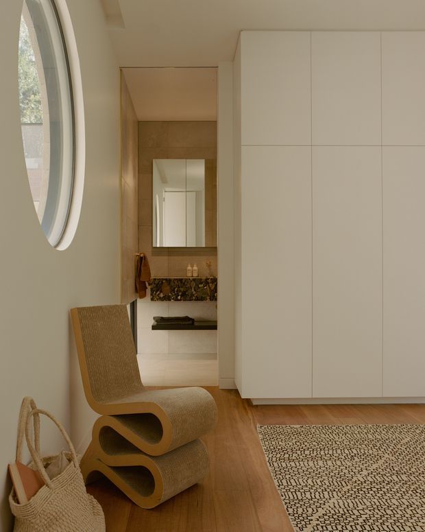 A warm minimalist interior hallway or entry area with a large curved oval window, pale built-in floor-to-ceiling cabinetry, timber flooring, and a sculptural woven lounge chair. A bathroom is visible through the opening, featuring tan wall tiles, a mirror, a stone or patterned vanity surface, and a wall-mounted basin. A textured neutral rug and woven basket add natural material accents.
