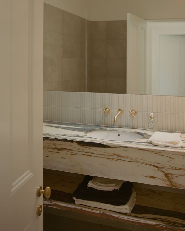A refined bathroom vanity viewed through a partially open white paneled door. The room features a wide marble vanity with prominent brown veining, an integrated white basin, brushed gold wall-mounted faucet and cross handles, a narrow vertically fluted tile splashback, large warm beige wall tiles, and open dark shelving below holding folded towels. A mirror reflects the tiled wall and doorway.