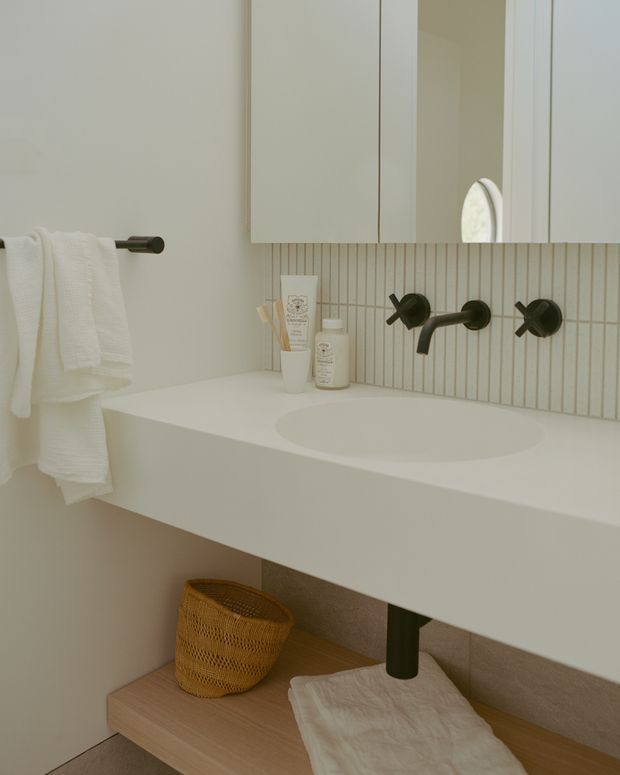 A softly lit contemporary bathroom vanity with a wall-mounted white basin and integrated countertop, matte dark wall-mounted tapware, pale vertical finger-tile splashback, mirrored upper cabinetry, and a light timber open shelf beneath. White towels and minimal toiletries add a calm, refined feel.