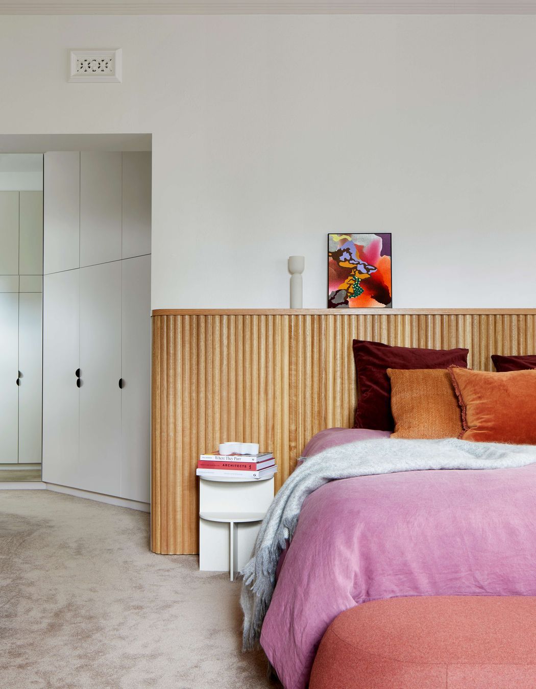 Master bedroom with headboard created with tasmanian oak wood dowels for a highly finished art deco minimalist feel. Cushions and bedding provide pops of colour in warm pink and jeweled colour oranges and burgundies. Pale pink  coloured carpet from Edel Carpets. Home located in Melbourne. See more from our Arch Deco Project.