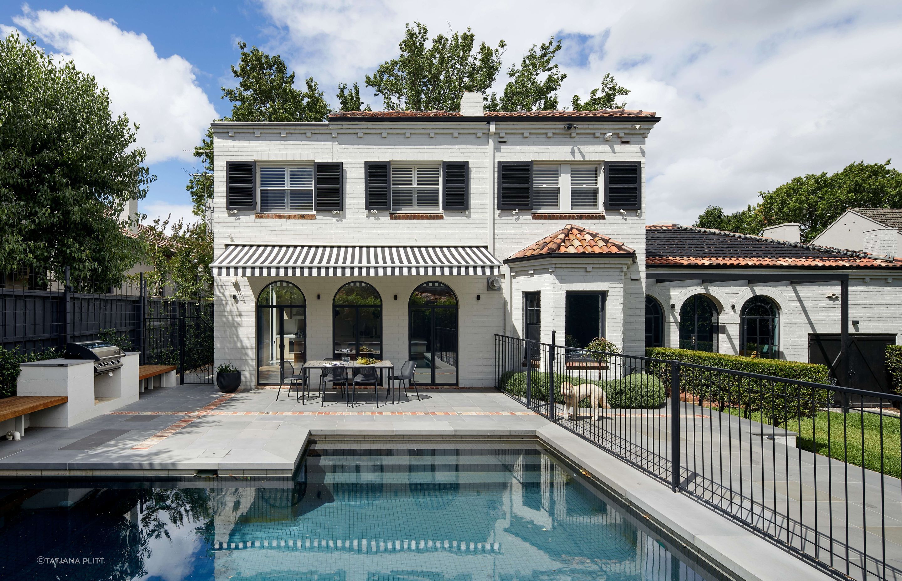 Art deco spanish mission house from the 1930’s renovated with updated paint with charcoal shutters. Rear space off kitchen features black and white striped awning, from Docril. See more from our Arch Deco Project.
