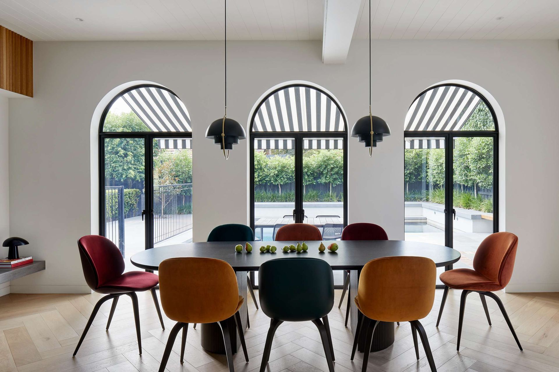 Modern art deco styled dining room featuring arched windows with black window frames. The room features pops of colour from the jewel coloured upholstered dining chairs from Gubi. The elliptical shaped black dining table with beveled half moon legs echo the curves of the art deco period. Overhanging black and brass pendant lights are from Gubi. The flooring is in pale oak in a herringbone pattern from Havwoods. Located in Melbourne, see more from our Arch Deco Project.