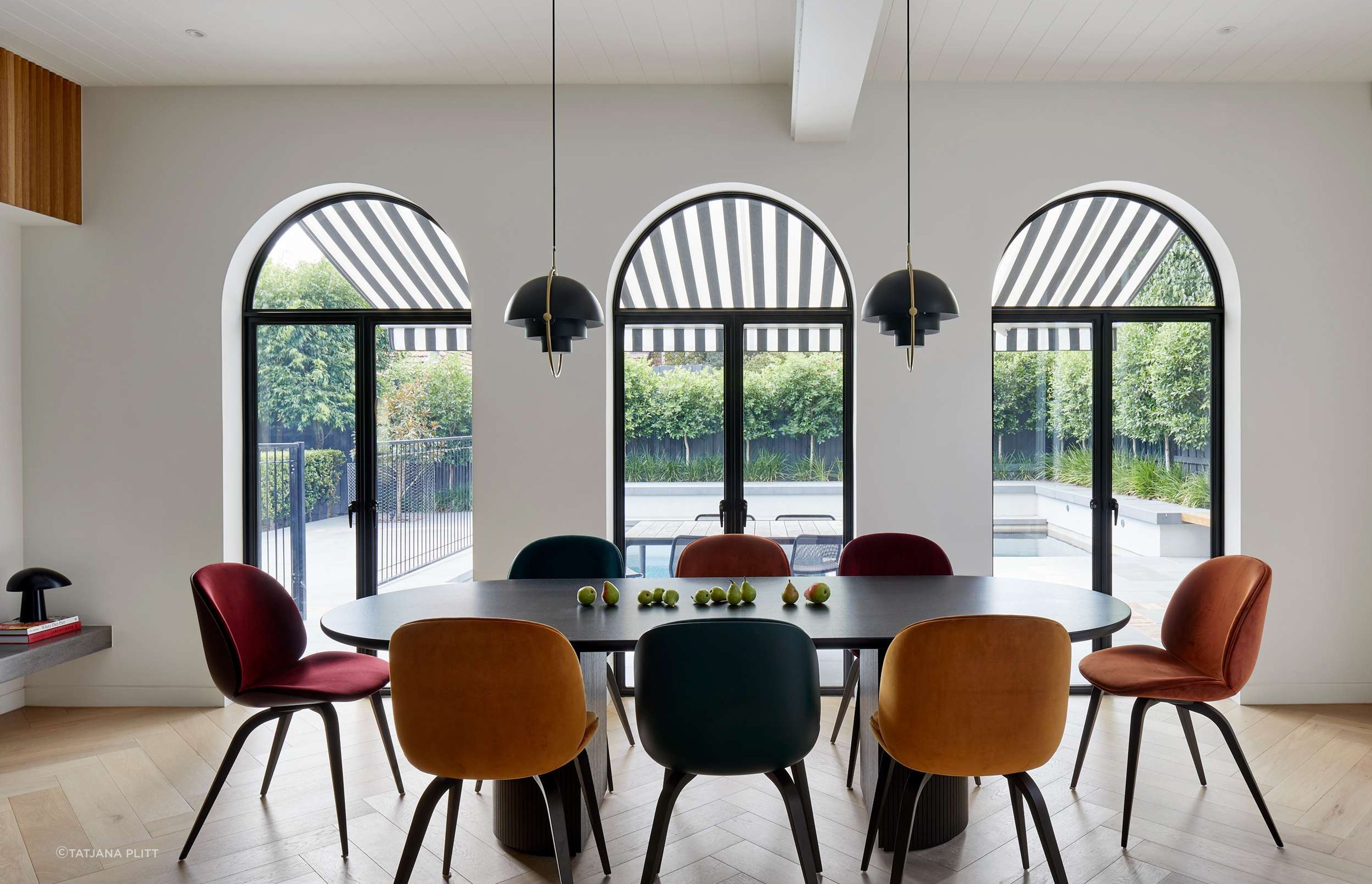 Modern art deco styled dining room featuring arched windows with black window frames. The room features pops of colour from the jewel coloured upholstered dining chairs from Gubi. The elliptical shaped black dining table with beveled half moon legs echo the curves of the art deco period. Overhanging black and brass pendant lights are from Gubi. The flooring is in pale oak in a herringbone pattern from Havwoods. Located in Melbourne, see more from our Arch Deco Project.