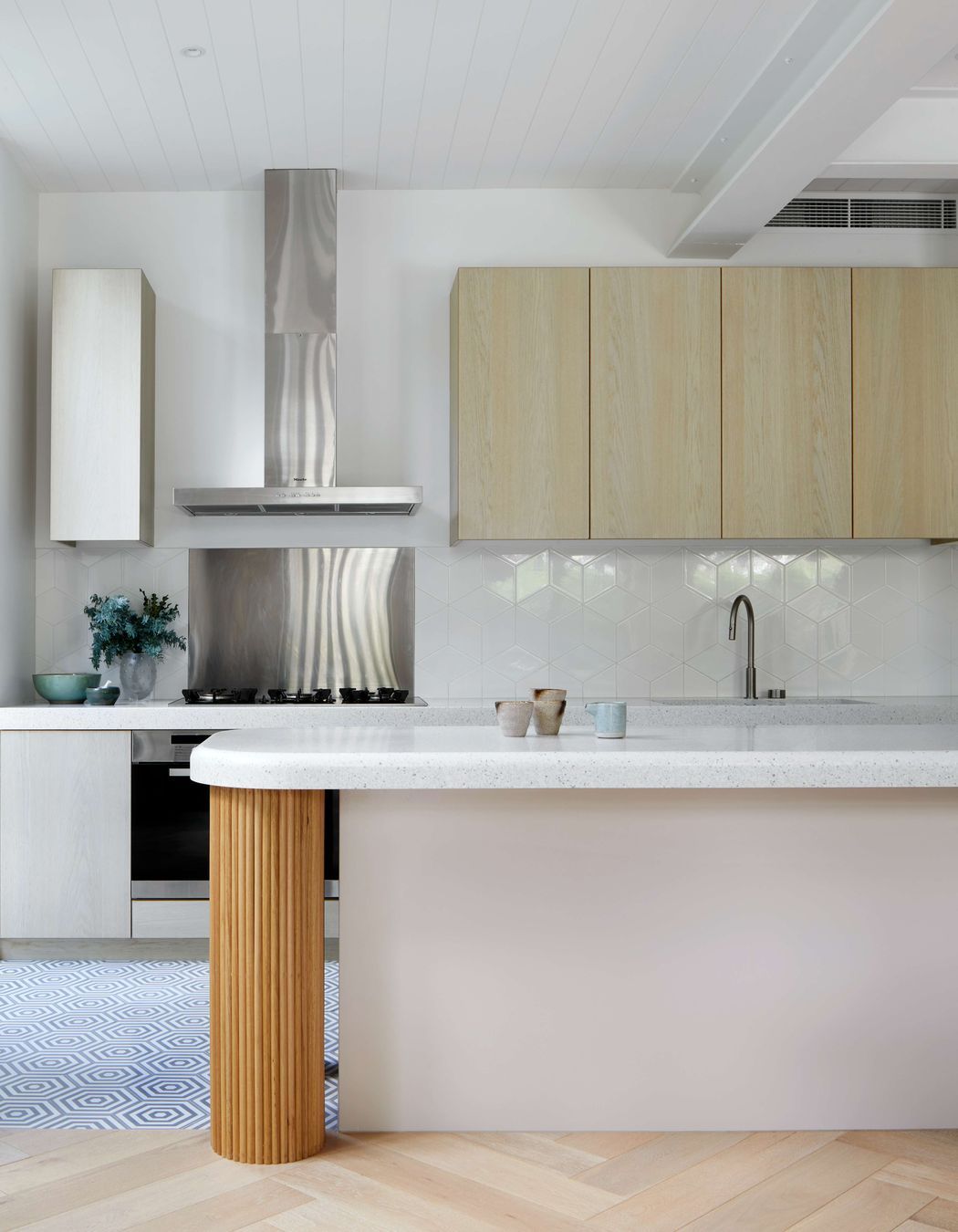 White kitchen with minimalist styling featuring white gloss diamond Perini tile backsplash, vibrant oak cool white veneer cabinetry, and a central cool grey cement hexagon patterned tiles on the floor, silver birch coloured corian stone bench top and oversized hand crafted pedestal legs using half round dowels as the finish for a contemporary and modern feel. Located in Melbourne, see more from our Arch Deco Project.