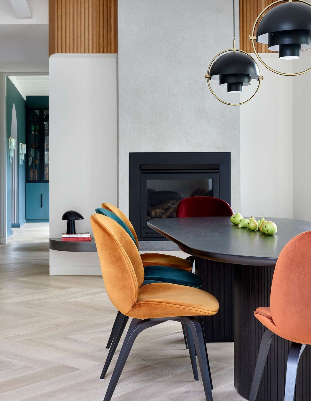 Modern inspired dining room with pops of colour from jewel coloured chairs to play off the art deco heritage of the home. The dining chairs are upholstered beetle dining chairs from Gubi, as is the elliptical shaped black dining table with bevelled half moon legs which reference simple design curves from the art deco period. Overhanging art deco inspired black and brass pendant lights are from Gubi. The flooring is in pale oak in a herringbone pattern from Havwoods, another nod to the art deco period of the home. Located in Melbourne, see more from our Arch Deco Project