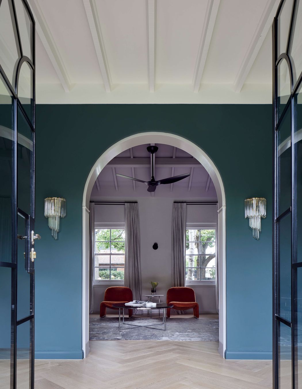 Library view from the living room through arched doorways and ceiling beams to keep with the art deco style of this house. Wall painted in dark green (Coriole) from Dulux (colour: Coriole). Crystal wall pendant lights frame the doorway and flooring is european oak laid in herringbone pattern with a matt lacquer for a minimalist styling.Project is a 1930s art deco spanish mission style house in Melbourne. See more from our Arch Deco Project.