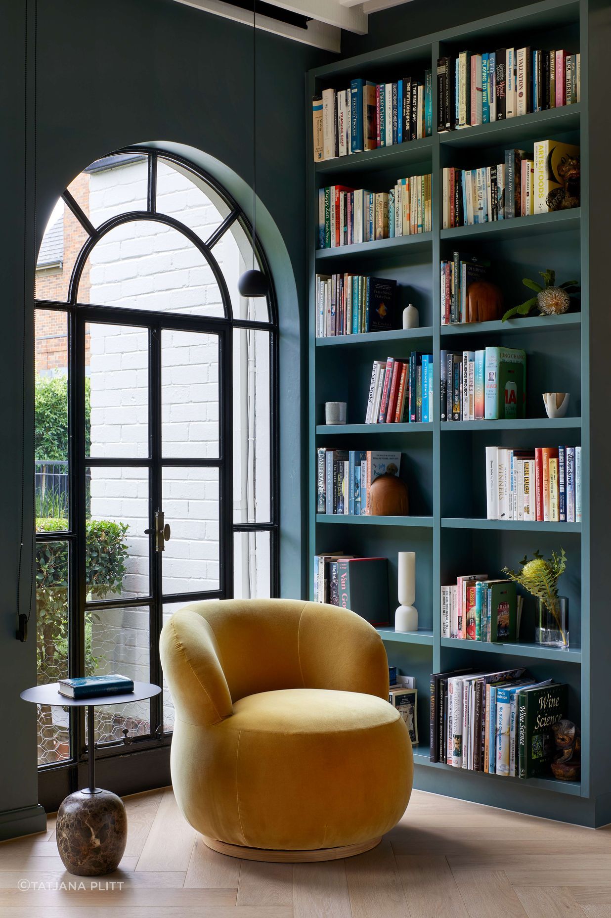 Art deco library featuring a large arched doorway framed by bookcases. Wall and bookcase painted in dark green from Dulux (colour: Coriole). Occasional Joy chair upholstered in elk velvet mustard from Jardan. Flooring in oak wood laid in herringbone pattern with a matt lacquer for a minimalist styling. The project is a 1930s art deco spanish mission style house in Melbourne. See more from our Arch Deco Project.