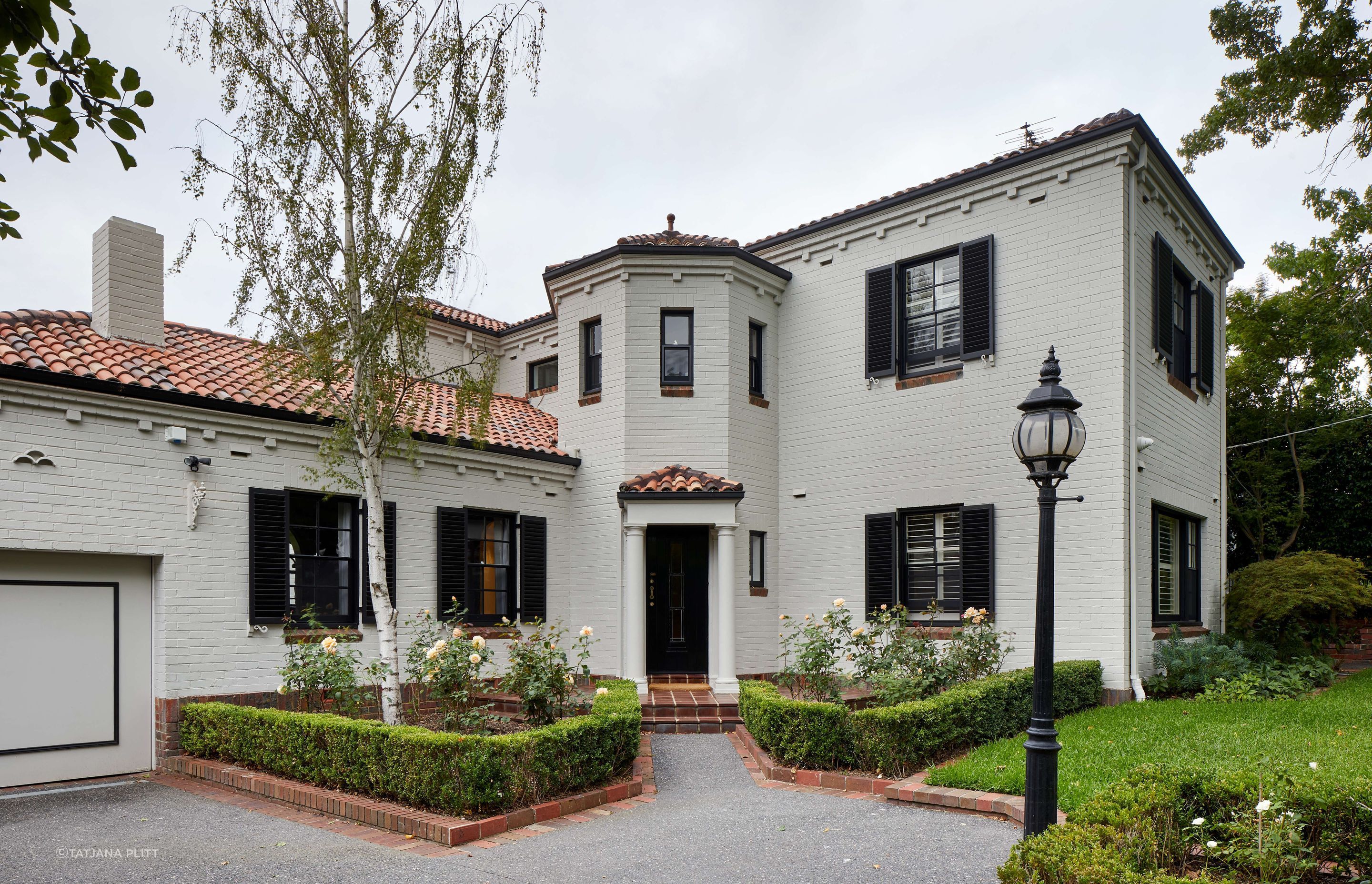 Renovated 1930’s art deco spanish mission style house in Melbourne. Art deco spanish mission house from the 1930’s renovated with updated paint with black shutters. Front of house with formal garden with box hedges and terracotta tiles. See more from our Arch Deco Project.