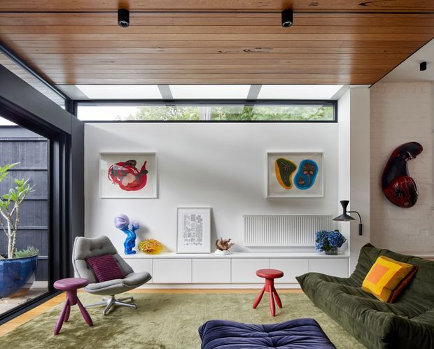 A bright, eclectic living room with a low white built-in media/storage unit, large black-framed glazing, and a long clerestory window. The room features a warm timber-lined ceiling with recessed spotlights, white-painted brick walls, sculptural artworks, a green sofa, grey lounge chair, colourful cushions, sculptural stools, and a large muted green rug.