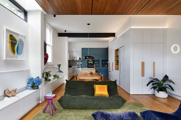 A bright, open-plan residential interior combining a living room, dining area and kitchen. The room features a deep green sofa, timber dining table, extensive white built-in cabinetry, blue kitchen joinery, timber flooring, and a distinctive warm timber slatted ceiling. Large windows provide natural light, while colourful artwork, plants and furnishings add character.
