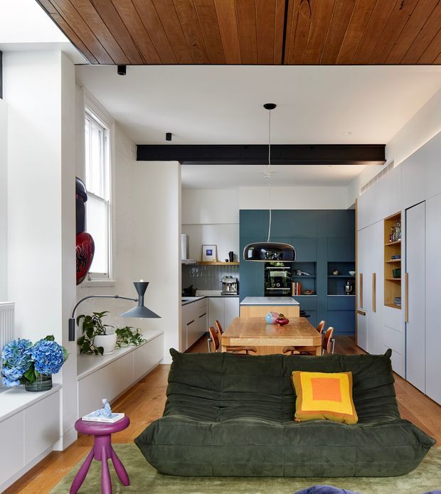 Bright open-plan living, dining and kitchen interior with a dark green sofa in the foreground, a timber dining table, and a compact blue built-in kitchen at the rear. White walls and cabinetry contrast with a warm timber ceiling and floor, while a dark exposed structural beam spans the room. Large windows, built-in shelving, plants, and colourful cushions add character to the contemporary space.