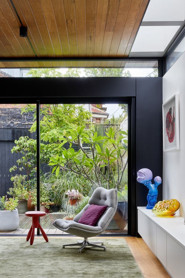 A stylish living room with a grey upholstered lounge chair and ottoman, a red accent cushion, and a small red side table. Full-height black-framed glazing opens views to a planted courtyard, while a warm timber-planked ceiling, dark feature wall, white built-in cabinetry, and skylight create a refined indoor-outdoor setting. Sculptural decorative objects and artwork add colourful accents.